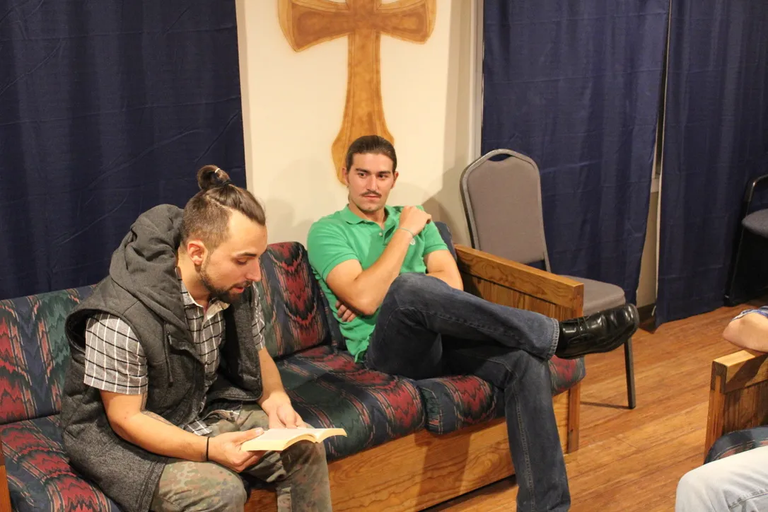 Two Teen Challenge residents reading the Bible together in a common area with a wooden cross on the wall