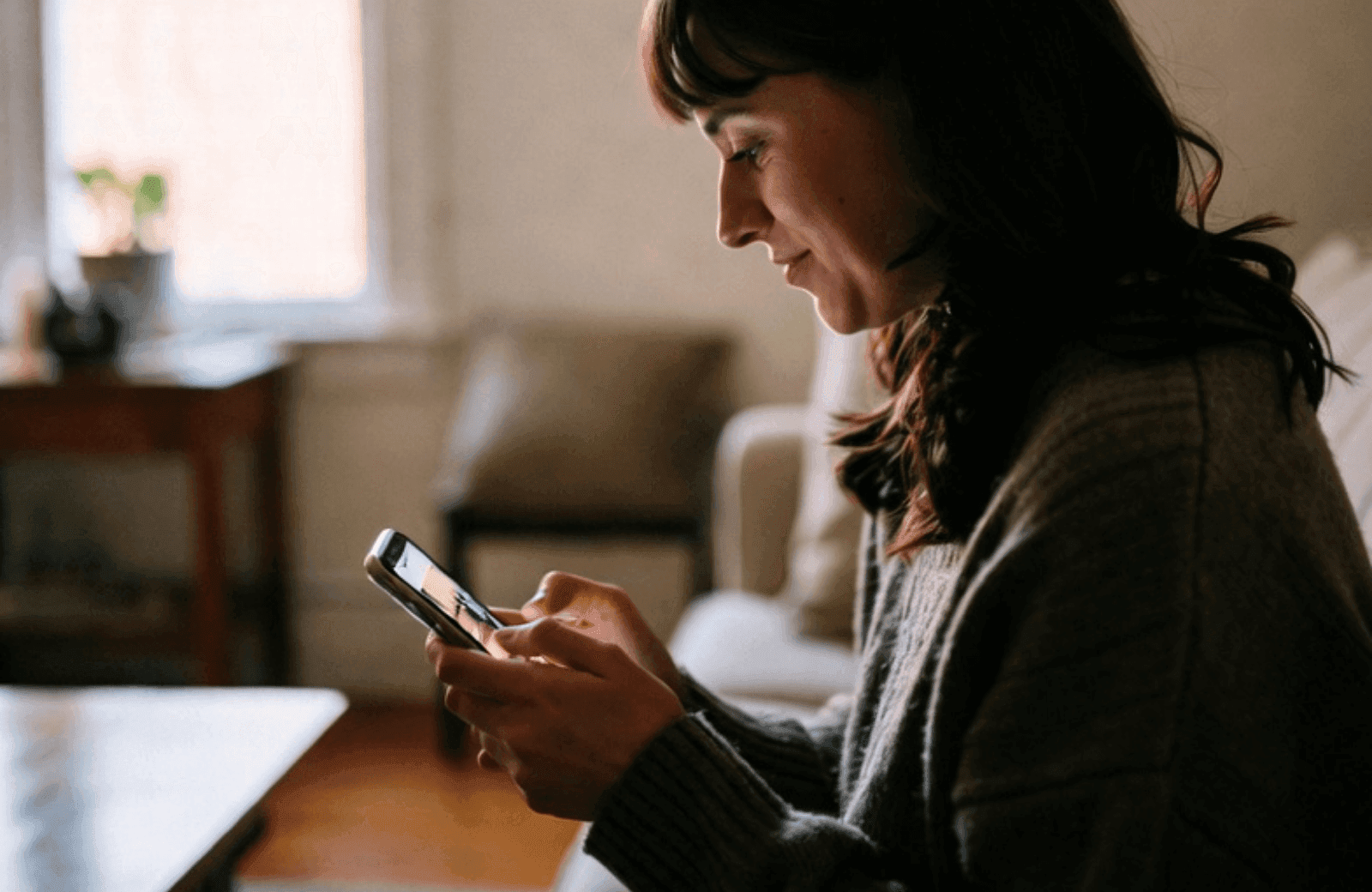 A thoughtful young woman sits on her couch in a warm Virginia living room, reading on her phone with a soft smile of realization, surrounded by family photos
