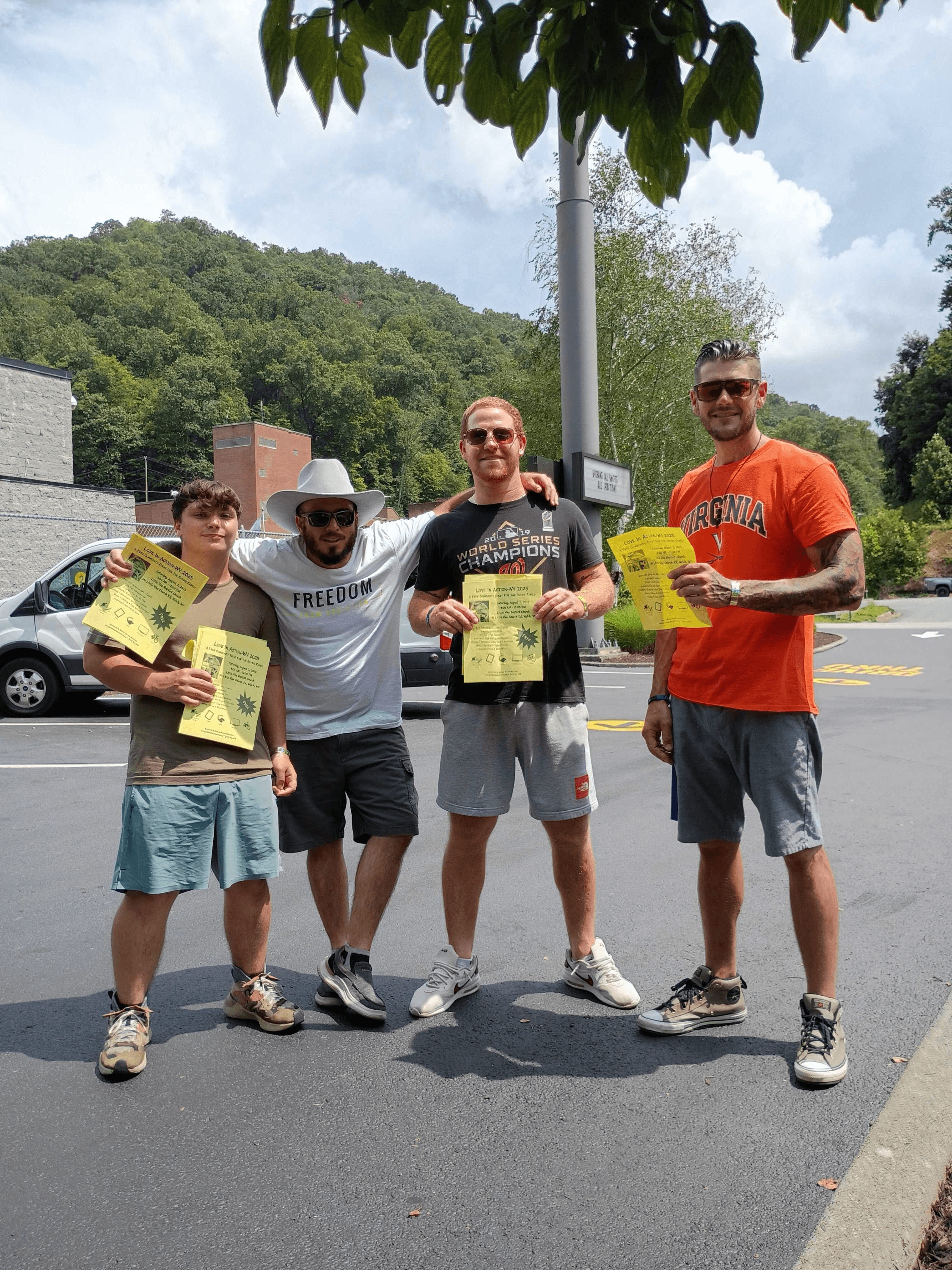 Four men standing in a parking lot in Welch, West Virginia holding yellow Love in Action 2025 outreach flyers, with Appalachian hills and a small town visible behind them.