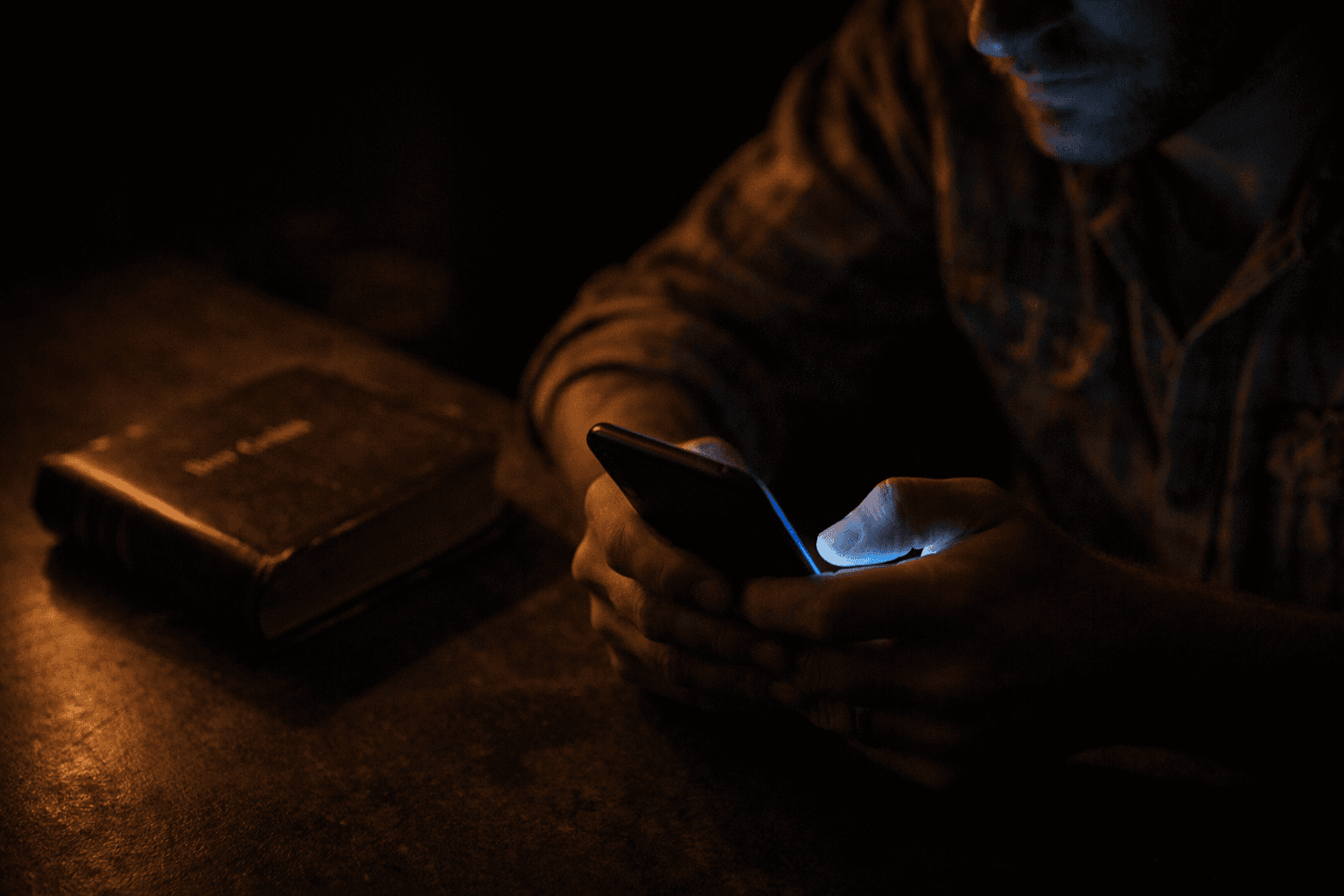 Hands holding a glowing smartphone in the dark while a Bible sits unused nearby