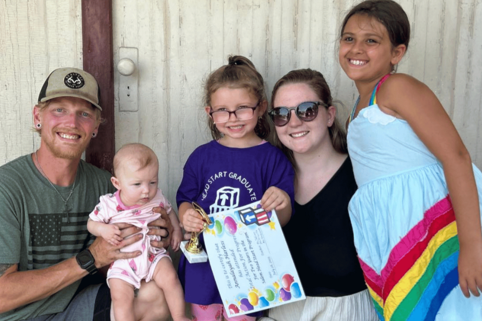 A smiling father holds his baby while sitting beside his wife and two daughters, celebrating their daughter’s graduation—evidence of life after addiction through Teen Challenge.